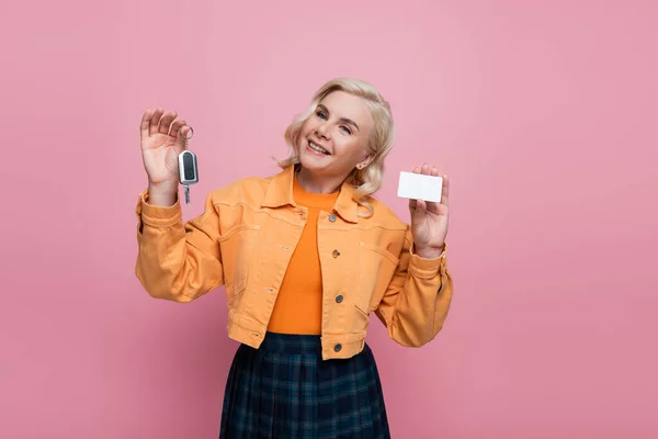 Blonde woman in jacket holding driving license and car key isolated on pink — стоковое фото