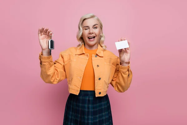 Blonde woman holding car key and driving license and winking at camera isolated on pink — стоковое фото