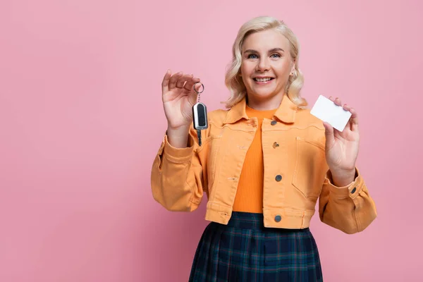 Happy blonde driver in jacket holding empty driving license and car key isolated on pink — Foto stock