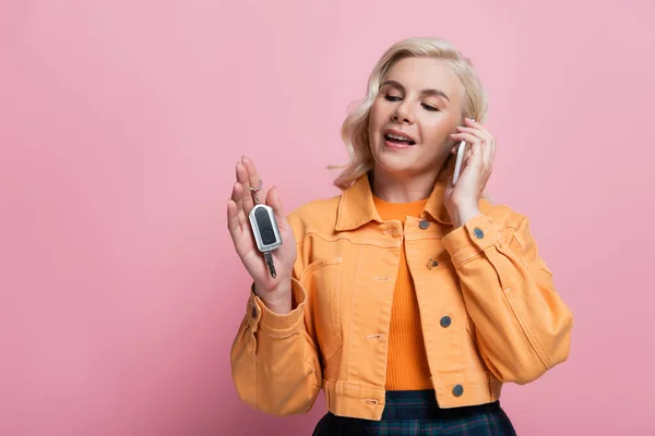 Blonde woman talking on smartphone and holding car key isolated on pink — Stock Photo