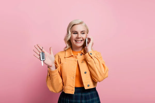 Blonde woman holding car key and talking on cellphone isolated on pink — Stock Photo