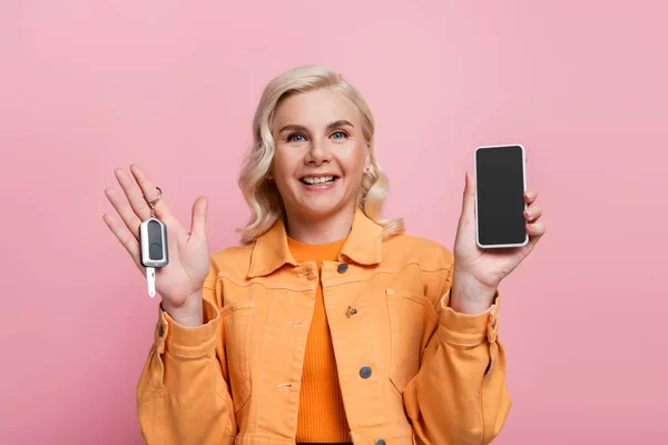 Весела жінка тримає смартфон і автомобільний ключ ізольовано на рожевому — стокове фото