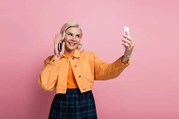 Smiling woman taking selfie on smartphone while holding car key isolated on pink - foto de stock