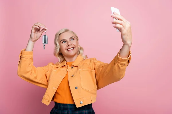 Cheerful woman holding car key and taking selfie on smartphone isolated on pink — Stock Photo