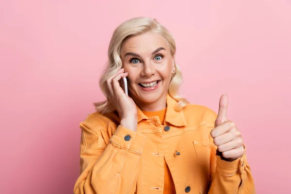 Blonde woman talking on smartphone and showing like gesture isolated on pink — стоковое фото