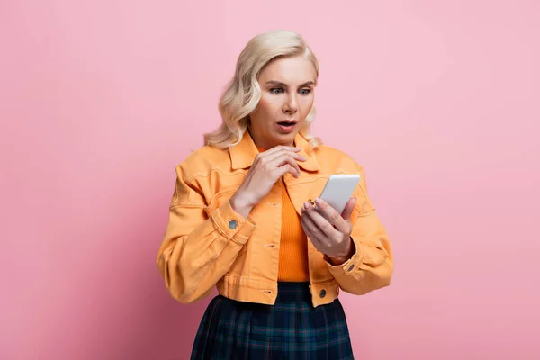 Shocked woman in orange jacket using smartphone isolated on pink — Foto stock