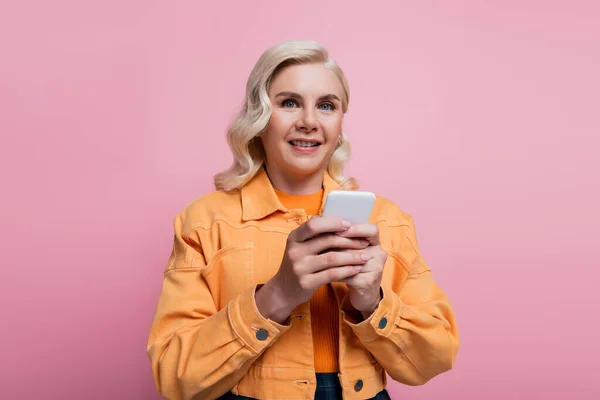 Усміхнена жінка в куртці тримає мобільний телефон і дивиться на камеру ізольовано на рожевому — стокове фото
