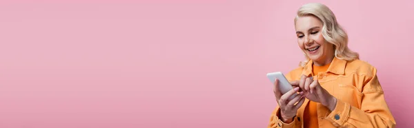 Blonde woman in orange jacket using smartphone isolated on pink, banner — Fotografia de Stock