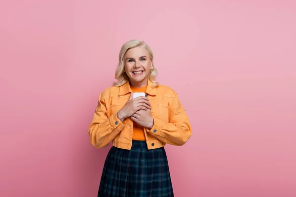 Happy blonde woman holding smartphone and looking at camera isolated on pink — стоковое фото