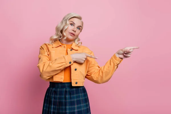 Blonde woman in jacket pointing with fingers isolated on pink — стоковое фото