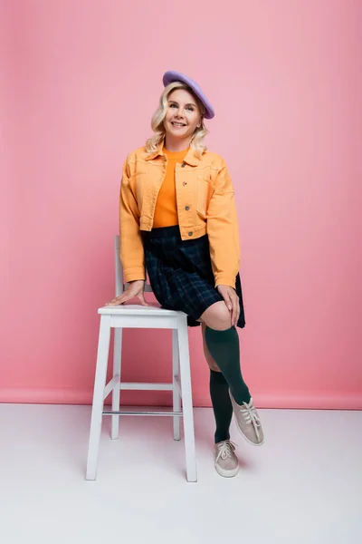 Smiling woman in skirt and beret posing near chair on pink background — Stock Photo