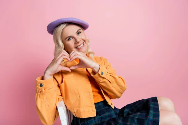 Smiling woman in beret showing heart gesture while sitting on chair isolated on pink — стоковое фото