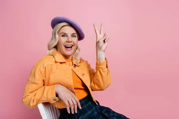 Positive woman in jacket and beret showing peace sign sitting on chair isolated on pink — стоковое фото