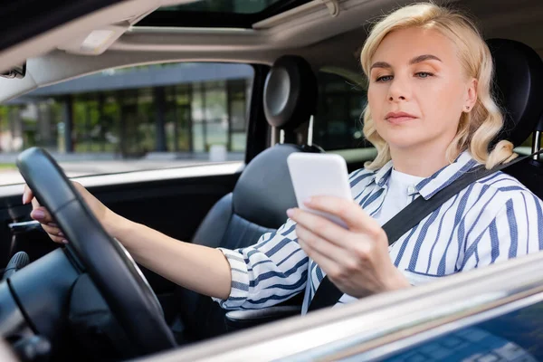 Blonde driving using smartphone while driving auto — стоковое фото