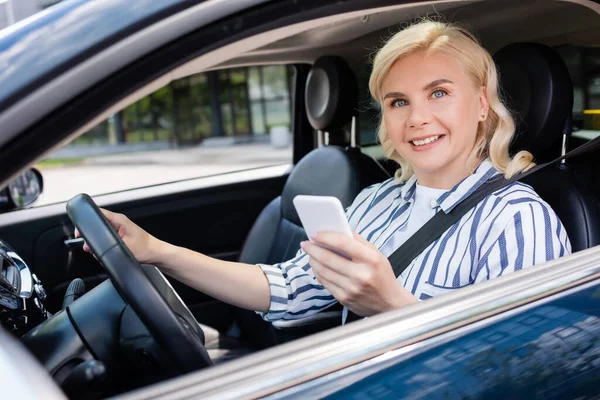 Blonde woman smiling at camera while sitting on driving seat in auto — Fotografia de Stock