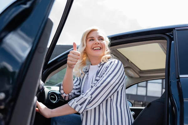 Low angle view of cheerful blonde woman showing thump up while opening door of car on urban street — Foto stock