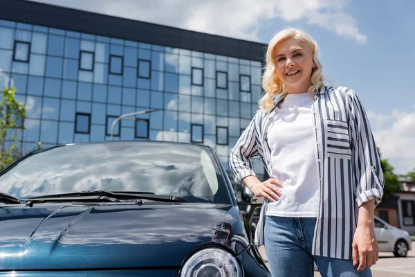 Positive woman holding hand on hip near car on urban street - foto de stock