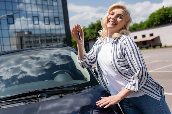 Positive blonde driver holding key near auto on urban street — Stockfoto