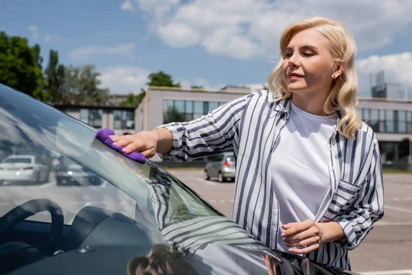 Blonde driver holding rag while cleaning windshield of car on urban street — Stock Photo