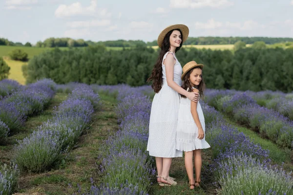 Full length of smiling mom and daughter in white dresses looking at camera in blooming field — Stockfoto