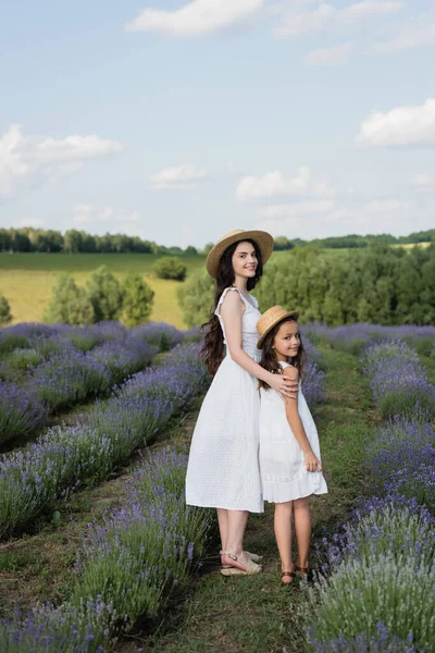Joyful mother and child in straw hats smiling at camera in lavender meadow - foto de stock