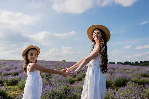 Cheerful woman and girl in straw hats holding hands and looking at camera in meadow — Foto stock