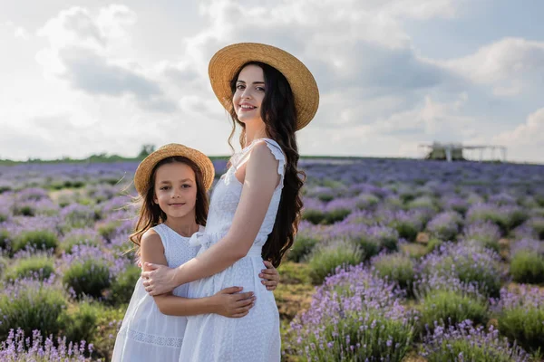 Happy woman and girl in white dresses hugging and looking at camera in field — Fotografia de Stock