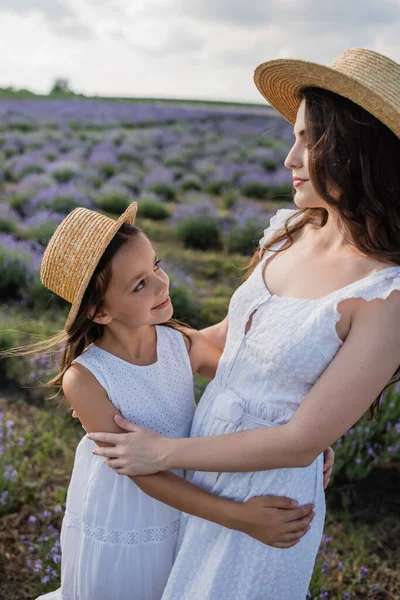 Mom and daughter in straw hats smiling at each other in flowering field - foto de stock