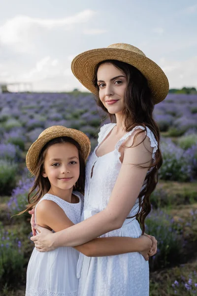 Brunette woman and daughter in straw hats hugging and looking at camera in lavender meadow — Stock Photo