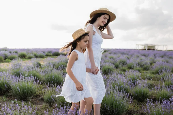 Woman and kid in straw hats holding hands while walking in lavender meadow — Fotografia de Stock