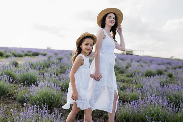 Smiling mom and daughter in straw hats holding hands and smiling at camera in lavender field - foto de stock