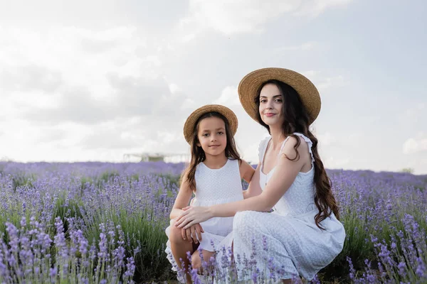 Mom and kid in straw hats and white dresses looking at camera near lavender in field — Photo de stock