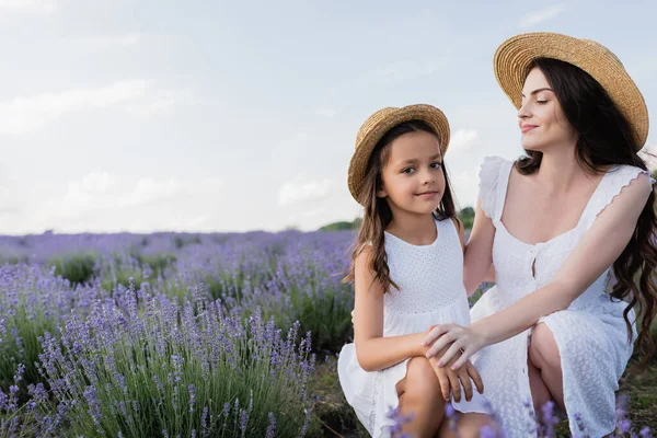 Girl in straw hat smiling at camera near happy mother in lavender meadow — Fotografia de Stock