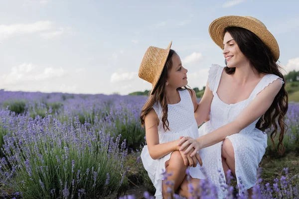 Жінка і дитина в білих сукнях і солом'яних капелюхах посміхаються один одному в квітучому лузі — стокове фото