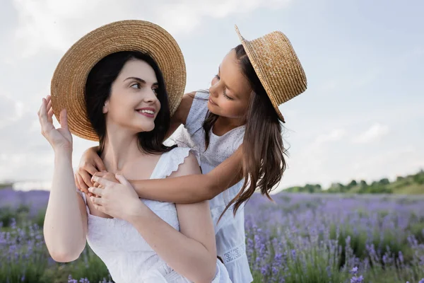 Child in straw hat embracing cheerful brunette mom in countryside — Photo de stock