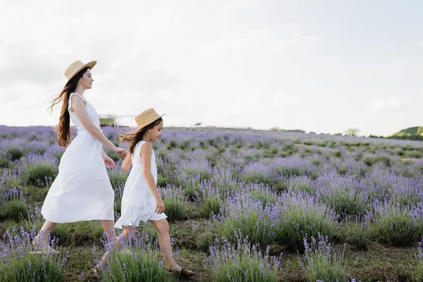 Side view of mom and child in white dresses and straw hats holding hands and walking in field - foto de stock