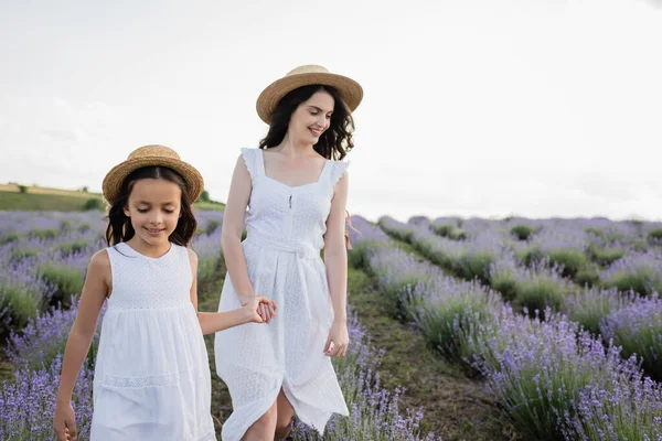 Brunette woman and girl in white dresses holding hands and walking in field - foto de stock