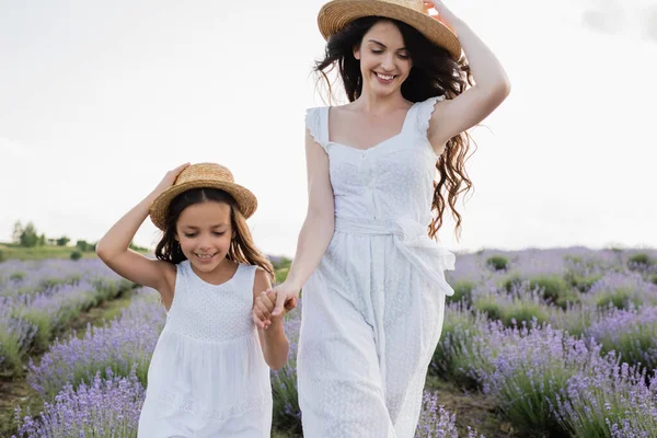 Smiling girl and woman in white dresses and straw hats holding hands in field — Stockfoto