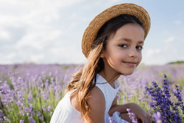 Задоволена дівчина в солом'яному капелюсі дивиться на камеру в полі з квітучим лавандою — стокове фото