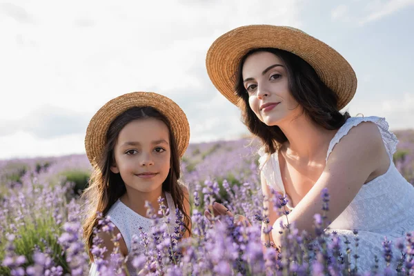 Мама и дочь в соломенных шляпах смотрят в камеру рядом с цветущей лавандой — стоковое фото