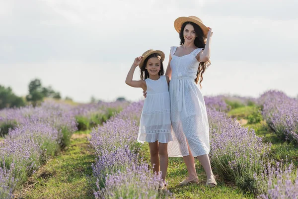 Cheerful mom and daughter in straw hats looking at camera in lavender meadow - foto de stock