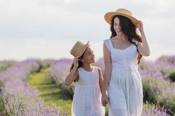 Joyful mother and girl in straw hats holding hands and looking at each other in field - foto de stock