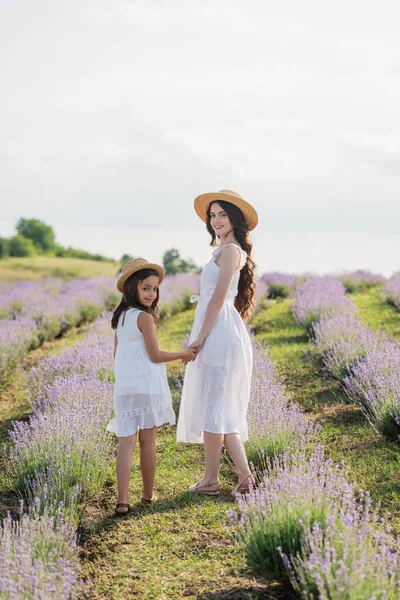 Brunette mom and daughter in white dresses holding hands and looking at camera in field - foto de stock