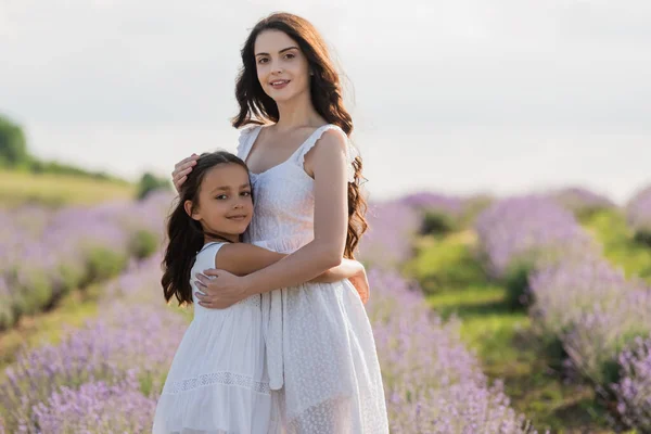 Girl and mom in white dresses embracing and looking at camera in blurred field — Photo de stock