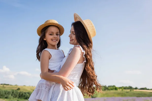 Cheerful girl in straw hat looking at camera in hands of mom outdoors — Foto stock