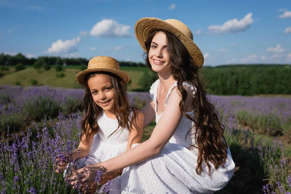 Happy brunette mom and daughter in straw hat smiling at camera in lavender field — Stock Photo