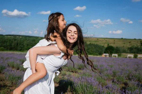 Cheerful brunette woman piggybacking daughter in flowering field — Foto stock