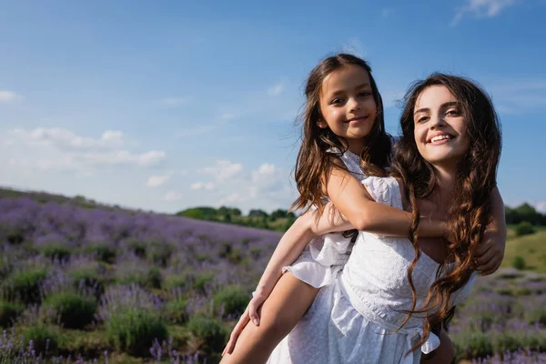 Brunette woman piggybacking daughter while having fun in field - foto de stock