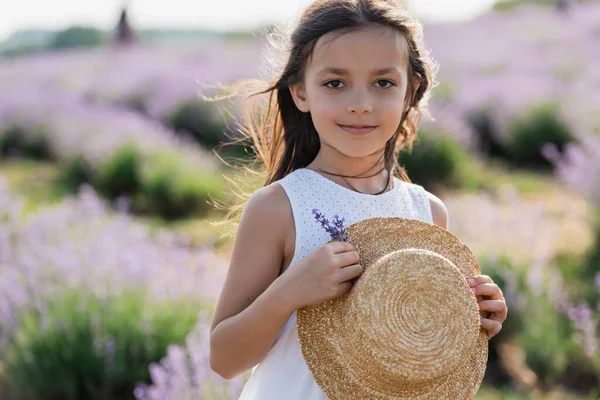 Girl with straw hat and lavender flowers looking at camera in blurred meadow — Photo de stock