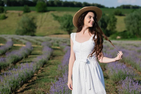 Positive woman in white dress and straw hat looking away in meadow with blooming lavender — Foto stock